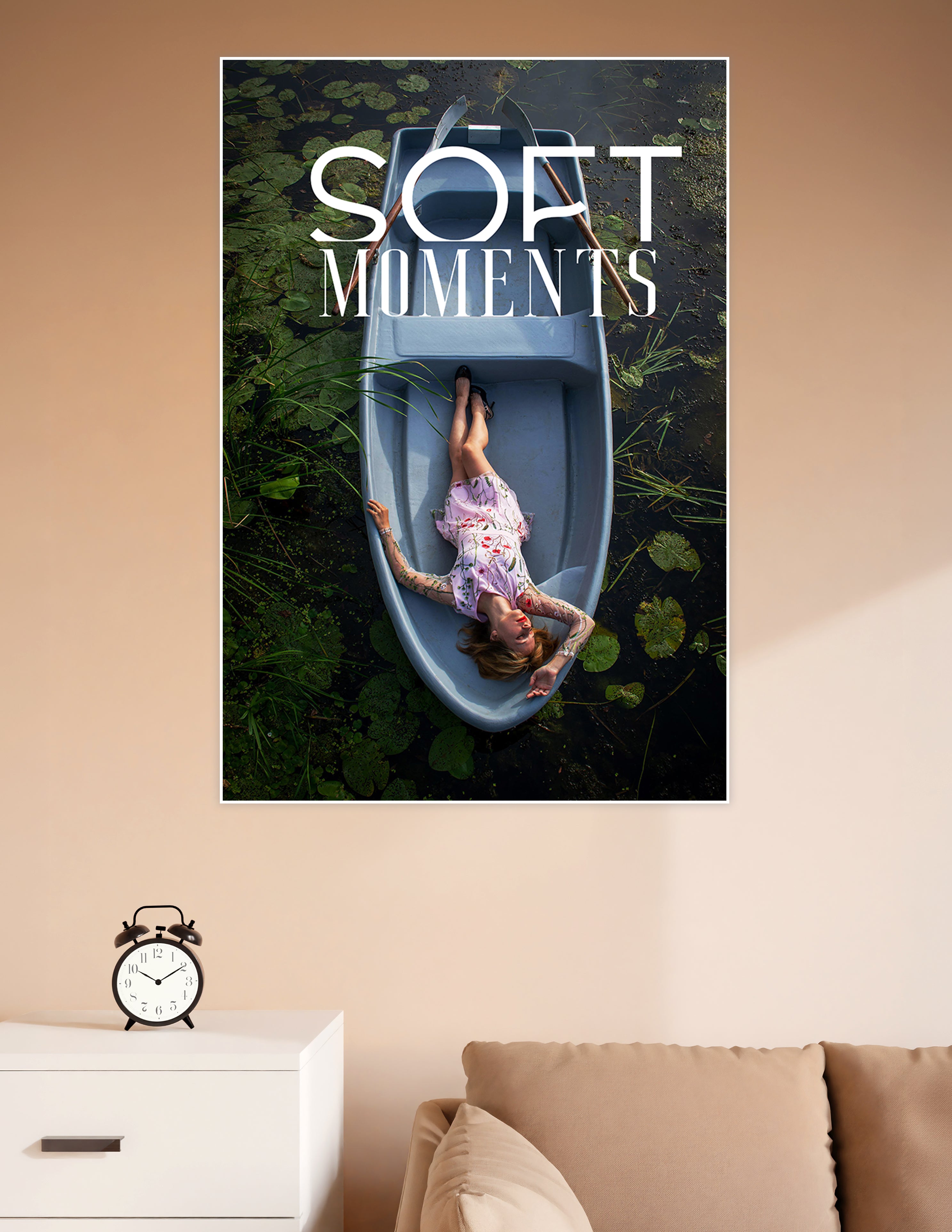 Aerial view of a woman in a floral dress lying in a pale blue rowboat surrounded by lily pads and dark water, titled Soft Moments