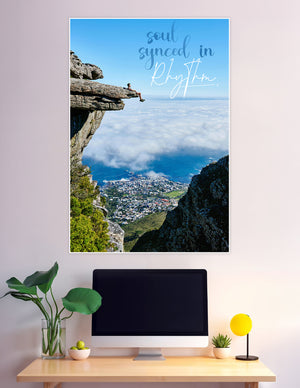 A person sitting on a rocky cliff edge high above clouds and a coastal city with the quote 'soul synced in rhythm' overlaid on a blue sky background