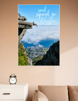 Person sitting on a rocky cliff overhang above clouds overlooking a coastal city with the quote soul synced in rhythm printed on the poster
